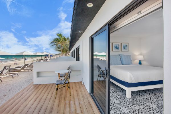 A beachfront room with a terrace, featuring a bed inside, and lounge chairs on the sand under umbrellas with a clear blue sky.