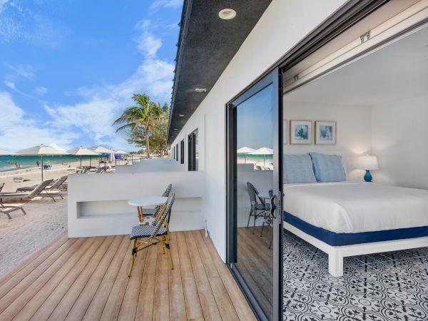 A beachfront room with a terrace, featuring a bed inside, and lounge chairs on the sand under umbrellas with a clear blue sky.