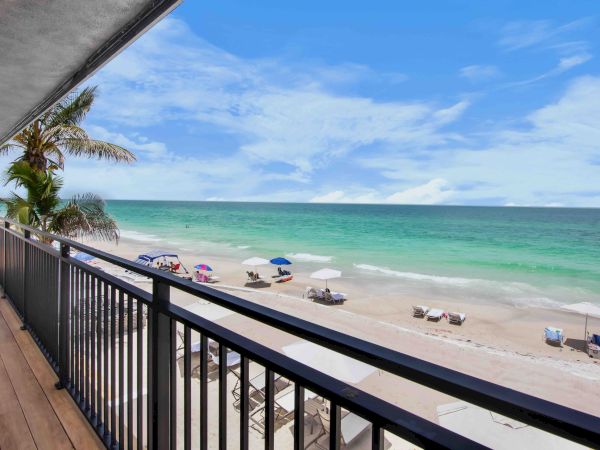 A sunny beachfront view from a balcony with palm trees, umbrellas and beach chairs along the sandy shore and turquoise ocean.