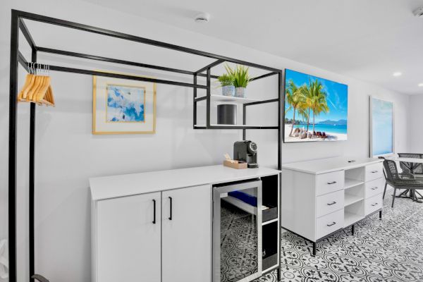 The image shows a modern room with a counter, coffee machine, framed art, TV, drawers, and a table with chairs on a patterned floor.