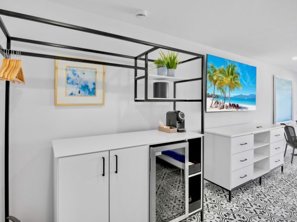 The image shows a modern room with a counter, coffee machine, framed art, TV, drawers, and a table with chairs on a patterned floor.