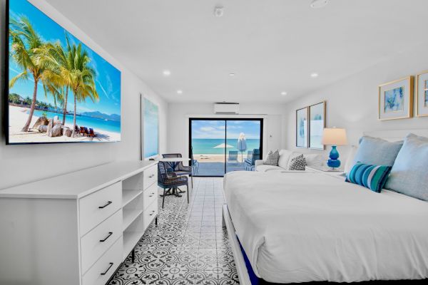 A bright bedroom with a large bed, beach artwork, a desk, and an ocean view through sliding glass doors, featuring decorative floor tiles.