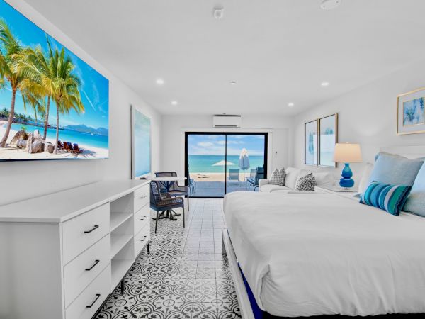 A bright bedroom with a large bed, beach artwork, a desk, and an ocean view through sliding glass doors, featuring decorative floor tiles.