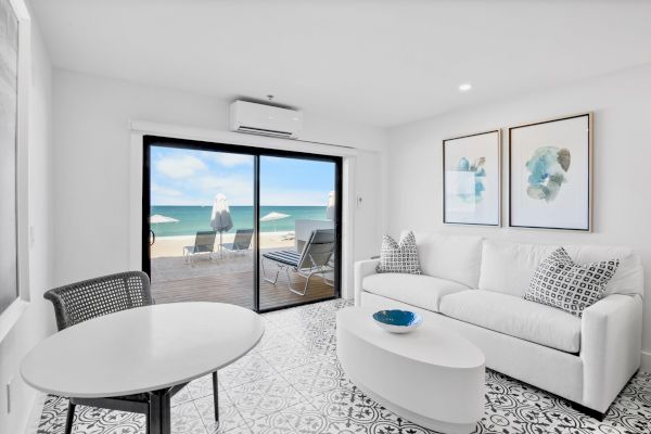 A bright living room with beach view, white furniture, and cozy décor; sliding glass doors lead to a patio with sun loungers.