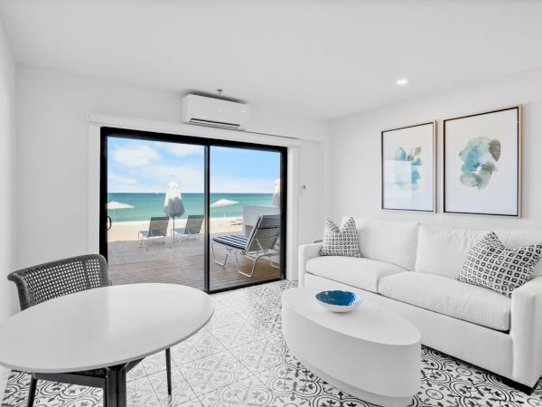 A bright living room with beach view, white furniture, and cozy décor; sliding glass doors lead to a patio with sun loungers.