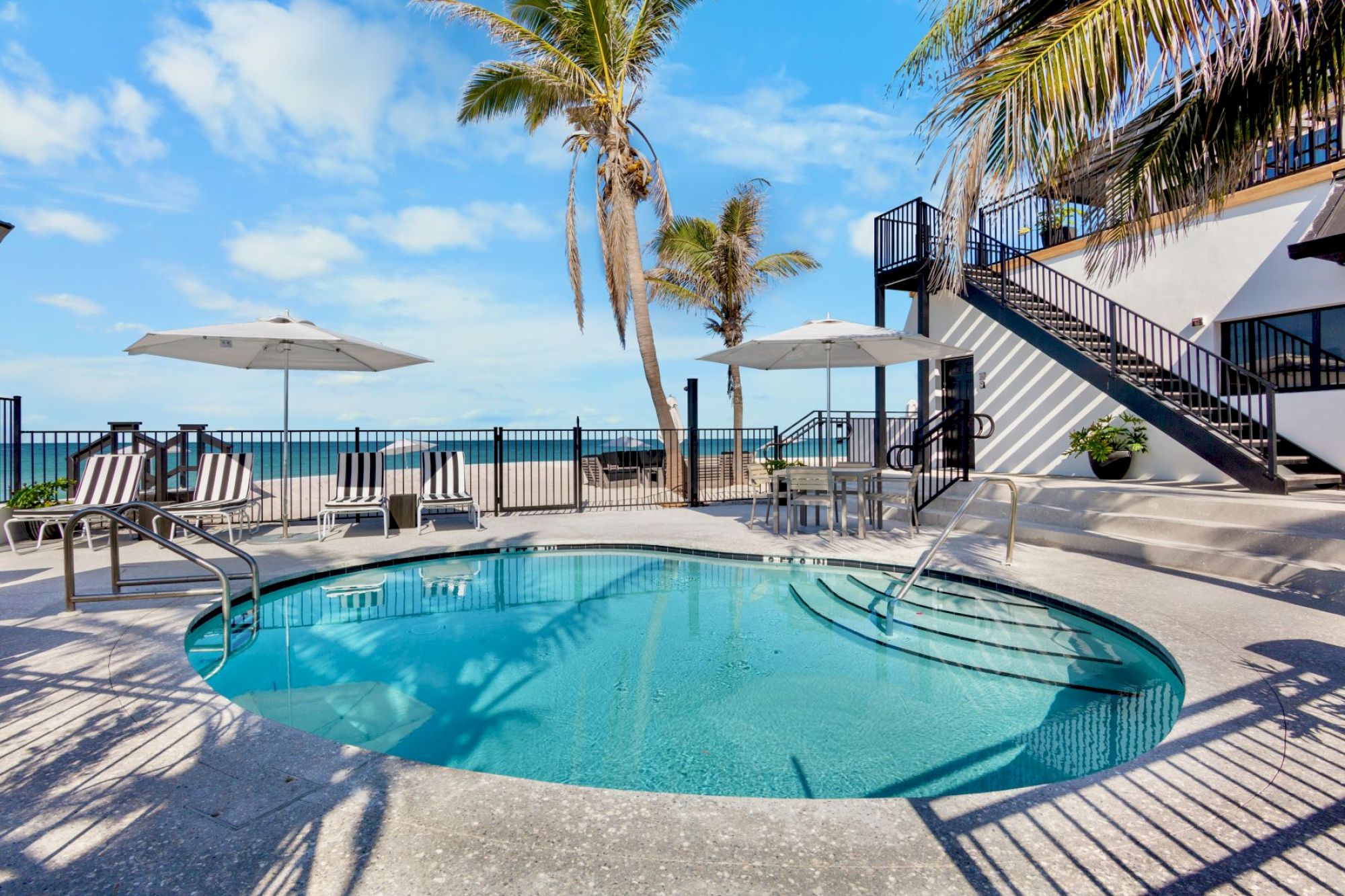 A circular pool with lounge chairs and umbrellas by the ocean, surrounded by palm trees and a staircase leading to an upper deck.