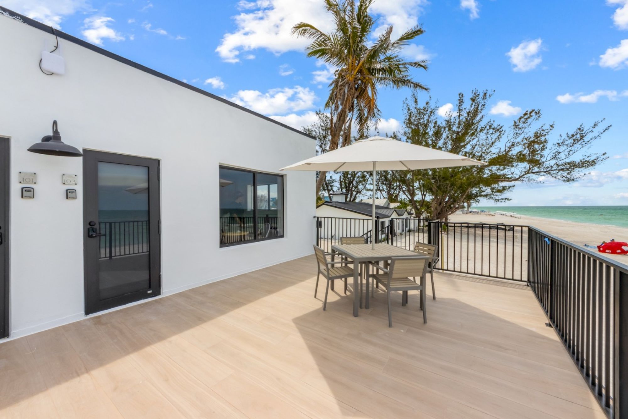 A beachfront patio with a white umbrella over a table and chairs, surrounded by railings, with palm trees and ocean in the background.
