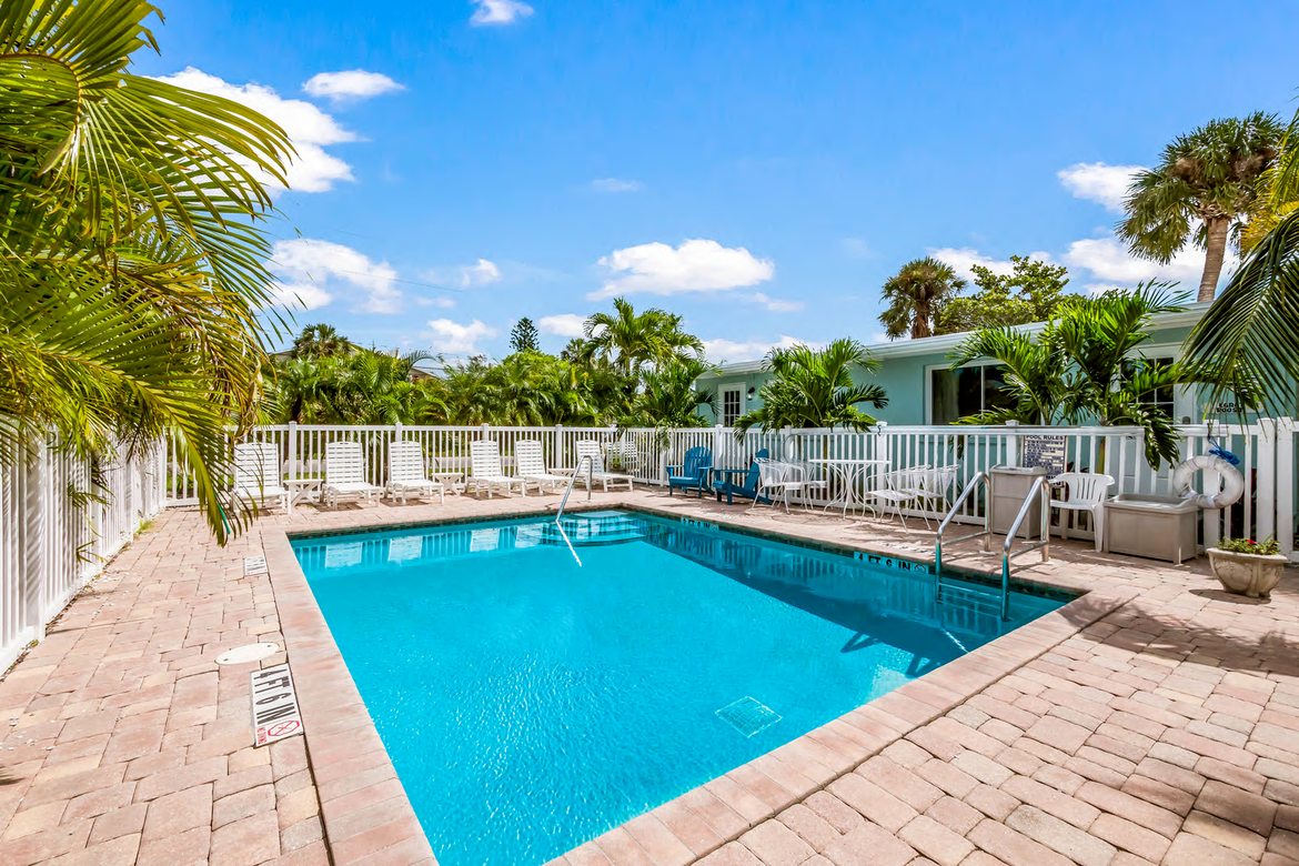A bright, sunny scene of a blue swimming pool surrounded by palm trees and lounge chairs with a clear sky overhead.