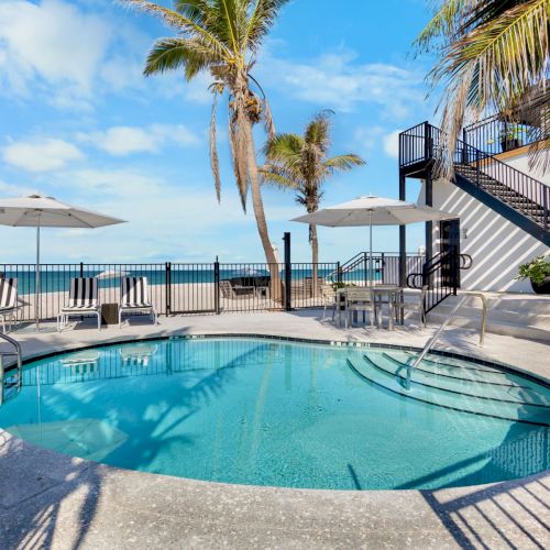 A small circular pool with a railing is surrounded by lounge chairs and umbrellas, near the ocean, with palm trees and a staircase nearby.