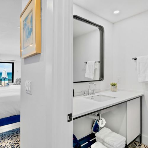 The image shows a modern beachfront hotel room with a bed, bathroom with a sink, mirror, towels, and a view of the ocean.