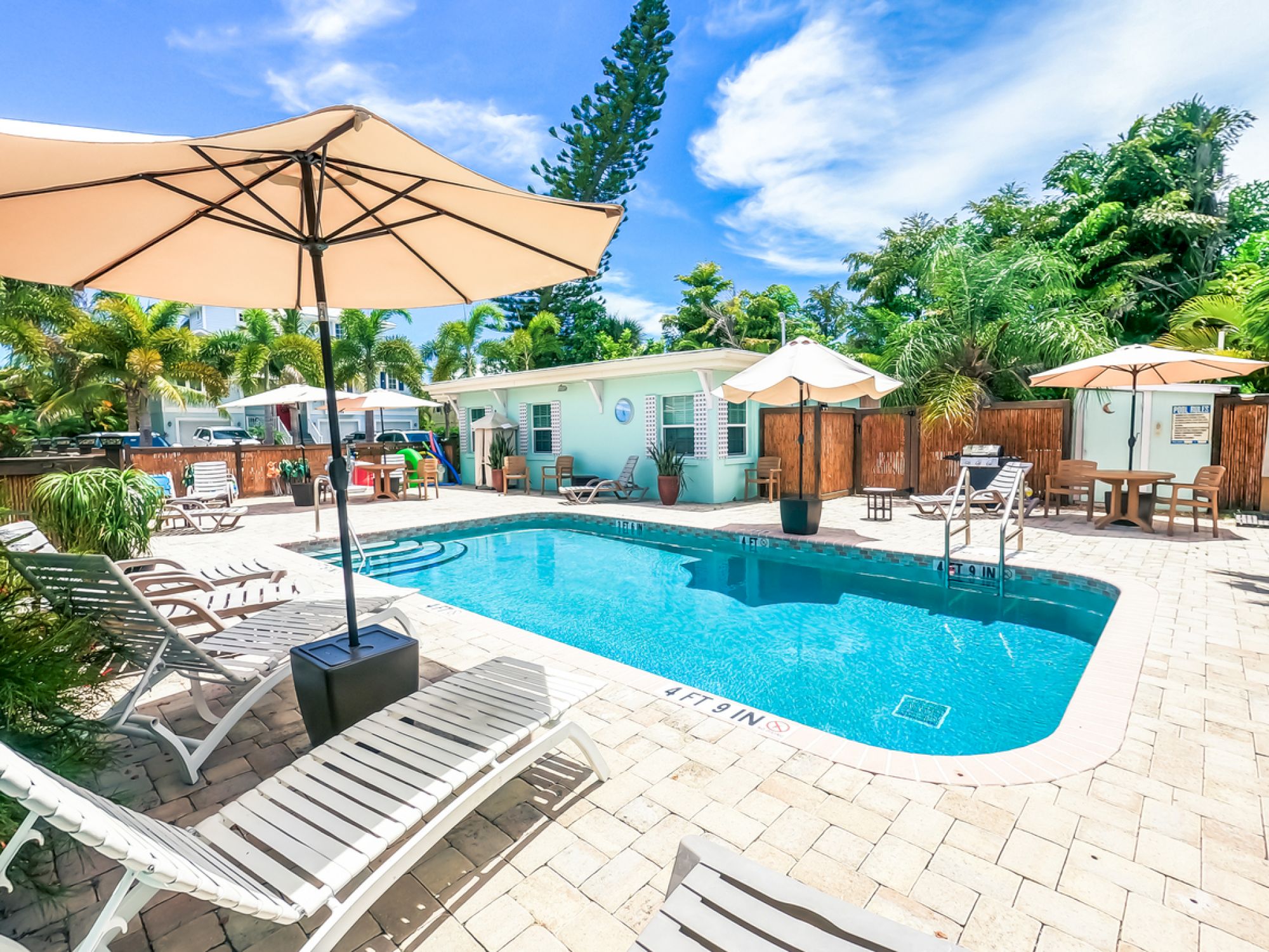 A sunny pool area with lounge chairs, umbrellas, and surrounding greenery, next to a small building.