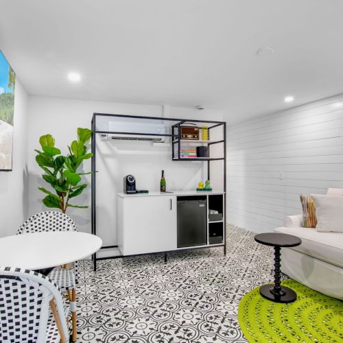 A modern room with patterned floor tiles, a white sofa, a small table, wall art, a TV, and a kitchenette with a plant nearby.