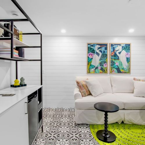 A cozy room with a white sofa, patterned tiles, two small tables, vibrant artwork, a countertop with items, and a bright rug.