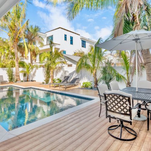 A luxurious outdoor pool area with lounge chairs, a patio set with an umbrella, surrounded by palm trees and modern buildings in the background.