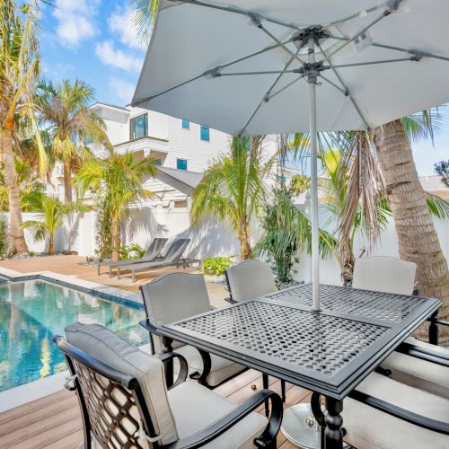 A patio table with chairs and umbrella by a pool, surrounded by palm trees and modern buildings under a blue sky.
