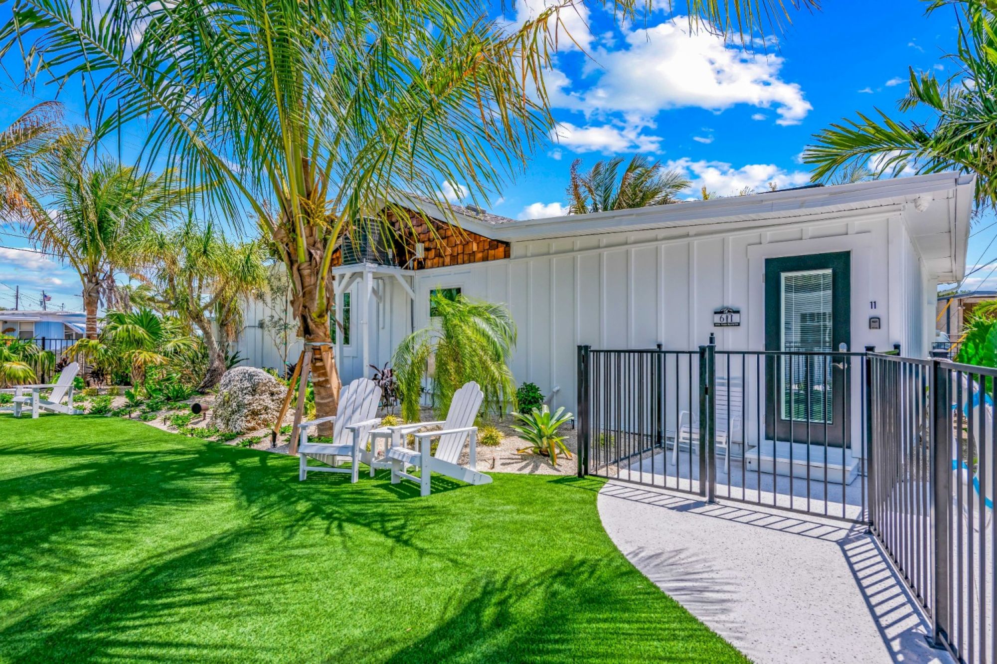 A modern house with white siding, lush greenery, palm trees, a lawn with outdoor seating, and a fenced entrance under a vibrant blue sky.