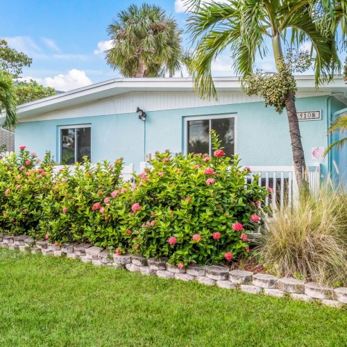 A light blue house with a white roof and trim, surrounded by palm trees and shrubs, with a well-kept lawn in the foreground.