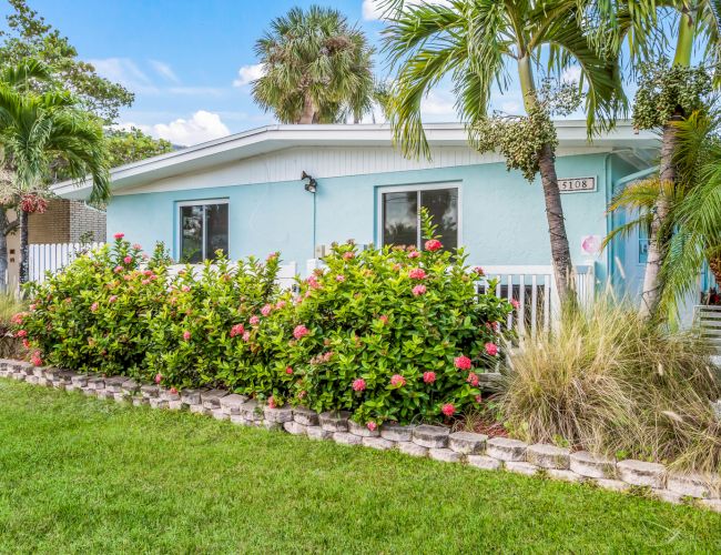 A light blue house with a white roof and trim, surrounded by palm trees and shrubs, with a well-kept lawn in the foreground.
