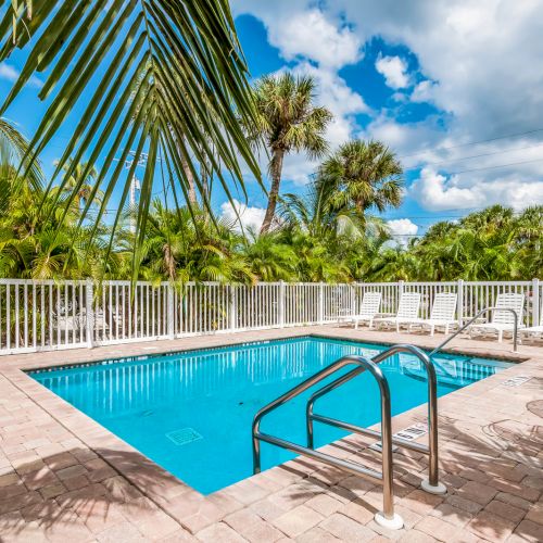 A small, rectangular swimming pool surrounded by a fence, palm trees, lounge chairs, and a table under a partly cloudy sky.