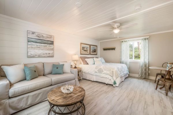 A cozy room with a sofa, bed, and wooden coffee table. Art on the wall and curtains frame the window. Light, airy decor with soft tones.
