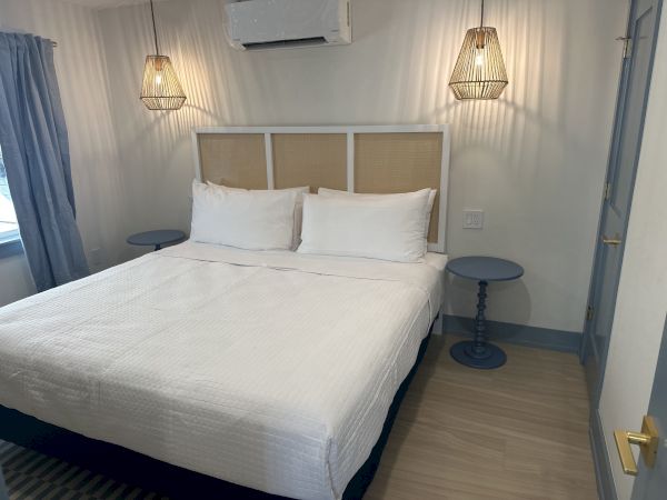 A neatly made bed with a white duvet, flanked by blue side tables, pendant lights, and a window with a curtain, all in a cozy bedroom.