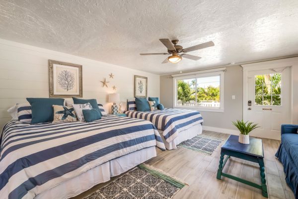 The image shows a bright bedroom with two striped beds, nautical decor, a ceiling fan, and a window with a view of greenery.
