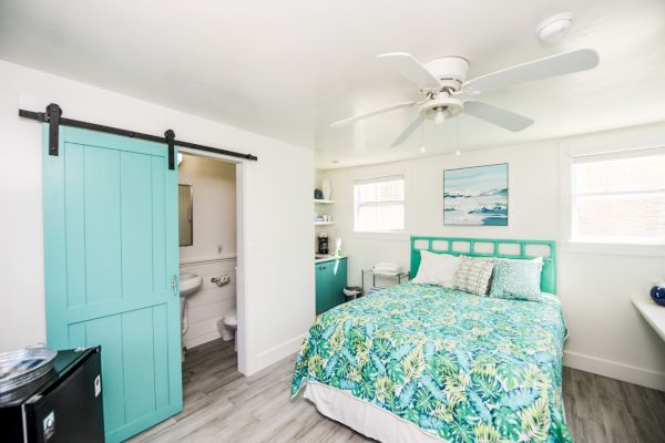 A bright, cozy bedroom with a teal sliding barn door, teal bed, floral bedding, ceiling fan, and an adjacent bathroom.