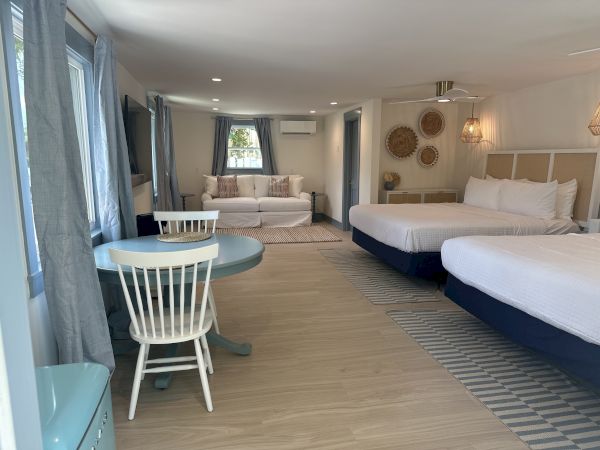 A spacious, well-lit room with two beds, a sofa, table with chairs, wall art, and windows, offering a comfortable and inviting atmosphere.