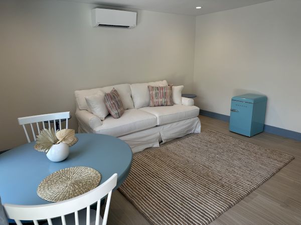 A cozy room with a white sofa, a round table and chairs, a small blue fridge, neutral walls, and a woven rug.