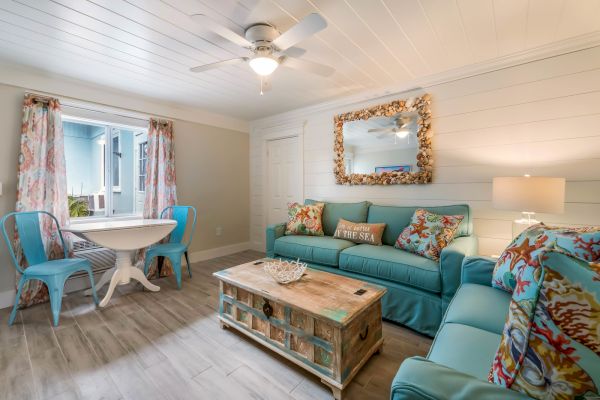 A cozy living room with blue sofas, a shell-themed mirror, decorative pillows, a rustic coffee table, and a small dining set by the window.