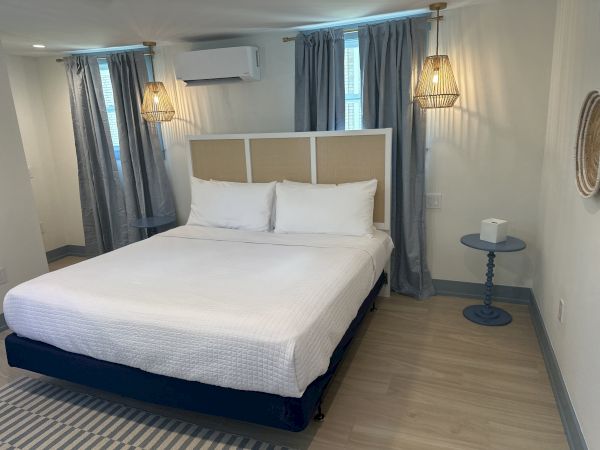 A neatly arranged bedroom with a large bed, two hanging lamps, gray curtains, a small round table, and a woven wall piece.
