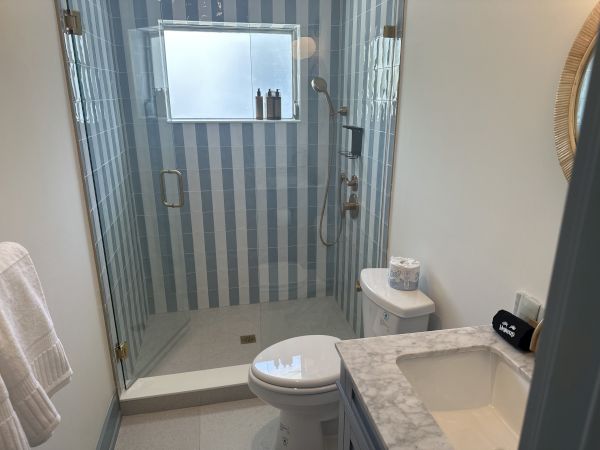 A bathroom featuring a glass shower with striped tiles, toilet, marble countertop sink, towels, and toiletries on a shelf completes the scene.