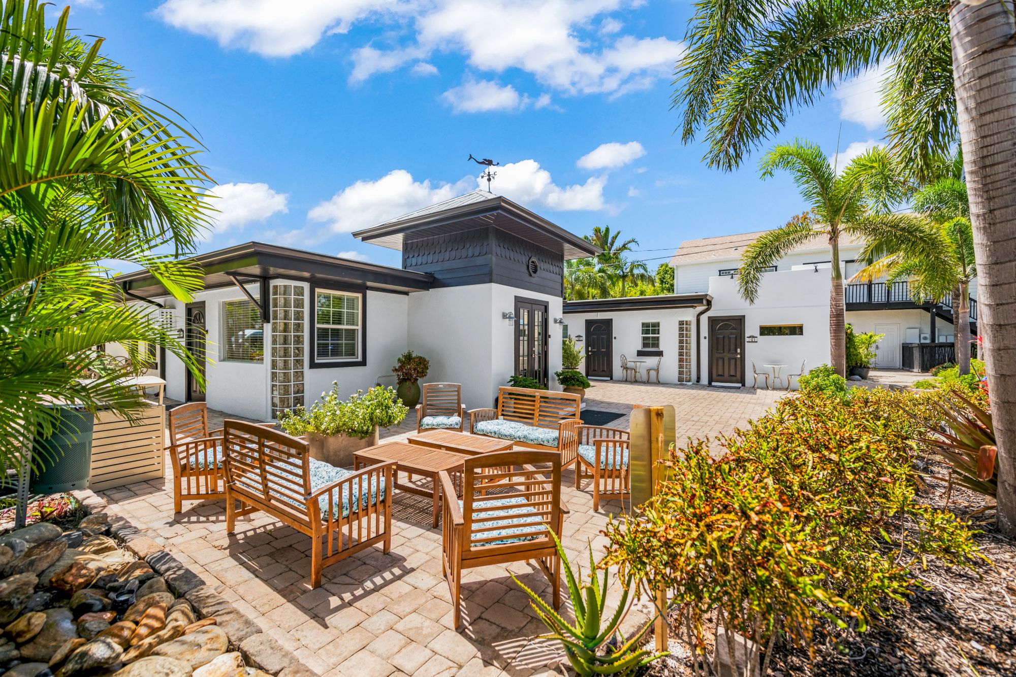 A sunny patio with wooden furniture, potted plants, and a white house surrounded by palm trees and a clear blue sky.
