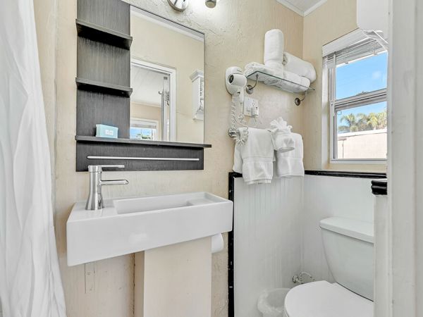 A small bathroom with a sink, mirror, wall shelves, towel rack, and window.
