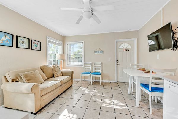 A cozy living room with a beige sofa, wall art, a small dining table, chairs, and a mounted TV, all under a ceiling fan.