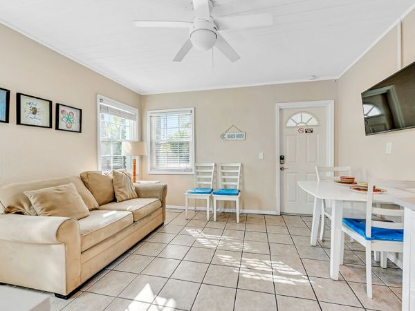 A cozy living room with a beige sofa, wall art, a small dining table, chairs, and a mounted TV, all under a ceiling fan.