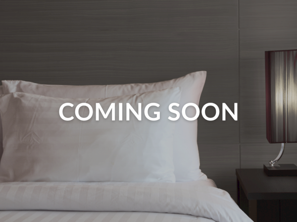 A bed with white pillows and sheets, a dark nightstand with a lamp, and the text "COMING SOON" overlaid on the image.