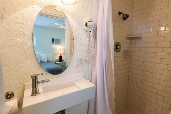 A bathroom with a white sink, oval mirror, shower with curtain, and hairdryer; bedroom visible in the mirror reflection.