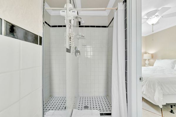 The image shows a bathroom with a white tiled shower, black accents, and a shower curtain near a bedroom with a bed and ceiling fan visible.