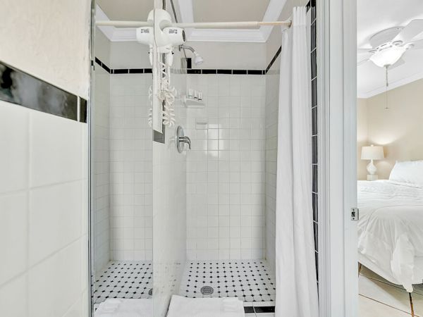 The image shows a bathroom with a white tiled shower, black accents, and a shower curtain near a bedroom with a bed and ceiling fan visible.