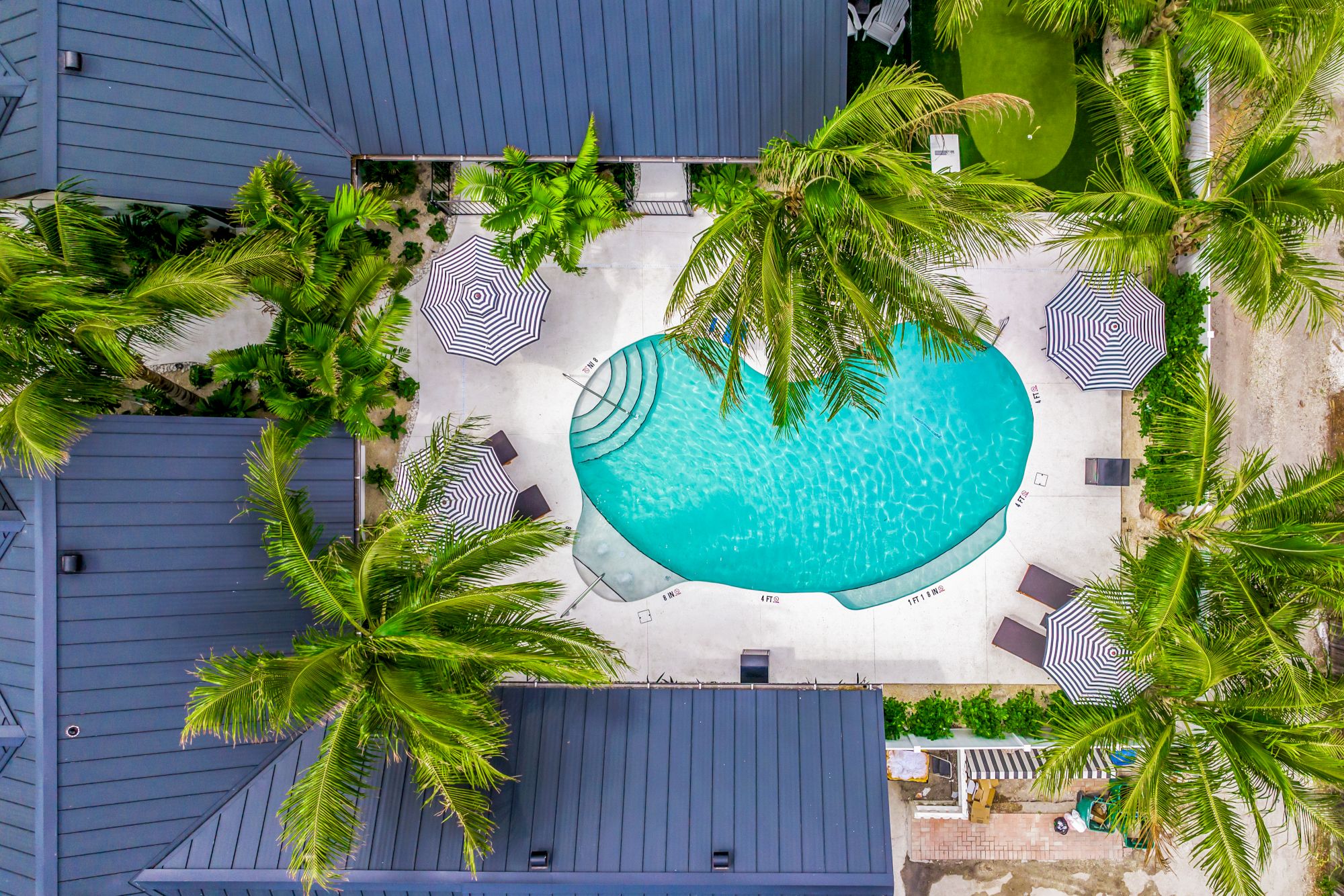 An aerial view of a kidney-shaped pool surrounded by palm trees, umbrellas, and lounge chairs, situated within a building complex.