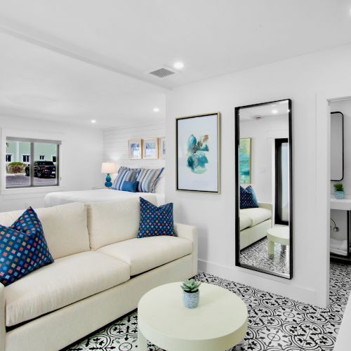 A modern living area with a white sofa, blue pillows, patterned floor, wall art, and a view into a small bathroom with a decorative touch.
