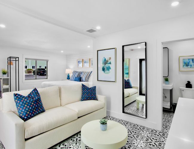 A modern living area with a white sofa, blue pillows, patterned floor, wall art, and a view into a small bathroom with a decorative touch.