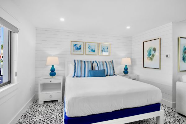 A bright bedroom with a white bed, striped pillows, blue accents, wall art, and decorative floor tiles.
