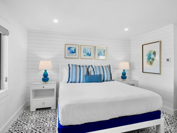 A bright bedroom with a white bed, striped pillows, blue accents, wall art, and decorative floor tiles.