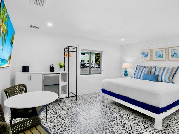 The image shows a modern bedroom with a double bed, patterned floor, a small table and chairs, and wall decorations, in a bright setting.