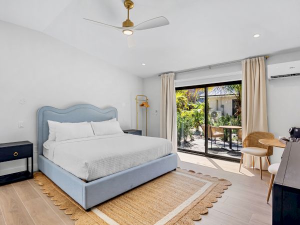 A bright, modern bedroom with a light blue upholstered bed, two black nightstands, a beige rug, and a balcony view through glass doors.