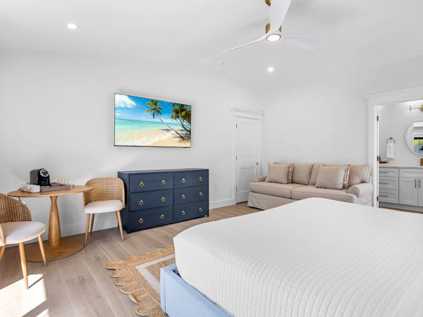 Bright hotel room with a king bed, beige sofa, navy dresser, wall TV, small desk with chairs, and a doorway to a bathroom in a sunny, airy space.