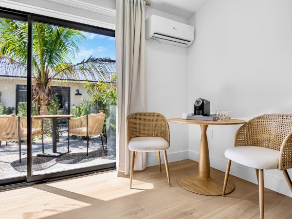A bright hotel room corner with a small round table and two wicker chairs, sliding glass doors to a sunny outdoor patio with palm trees.