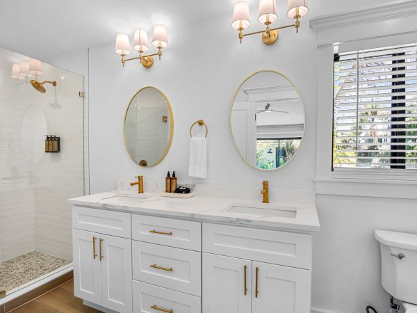 A bright, white bathroom with a double sink vanity, gold hardware, two oval mirrors, wall sconces, a glass shower, and a white toilet by a window.